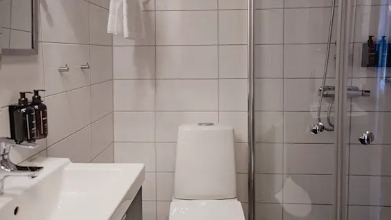 Ett rent och modernt hotellbadrum med vita kakelplattor, med handfat, toalett och en dusch med glasvägg.