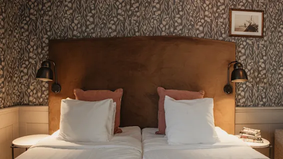 A cozy hotel room with two beds, a brown headboard, and patterned wallpaper. Bedside lamps and a framed picture complete the scene.