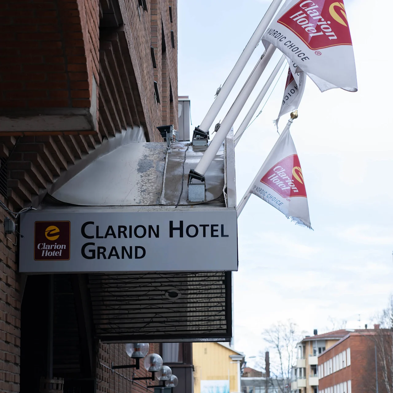 Skylt för "Clarion Hotel Grand" hänger på en tegelfasadsom med flaggor. Gatubelysning och byggnader i urban bakgrund. Flaggor med texten "Clarion Hotel" och "Nordic Choice".
