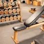 Træningsbænk placeret på et gummi-gymgulv; i nærheden holder en træreol forskellige håndvægte, hvilket tyder på et fitnessstudie-miljø med yderligere udstyr i baggrunden.