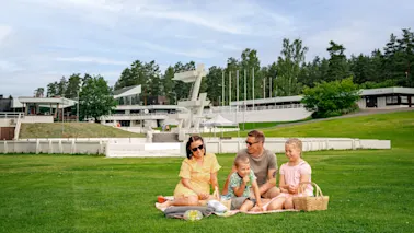 Perhe istuu piknikhuovalla, syö ja keskustelee. Heitä ympäröi laaja vihreä nurmikko, jossa on puita ja moderneja rakennuksia taustalla.