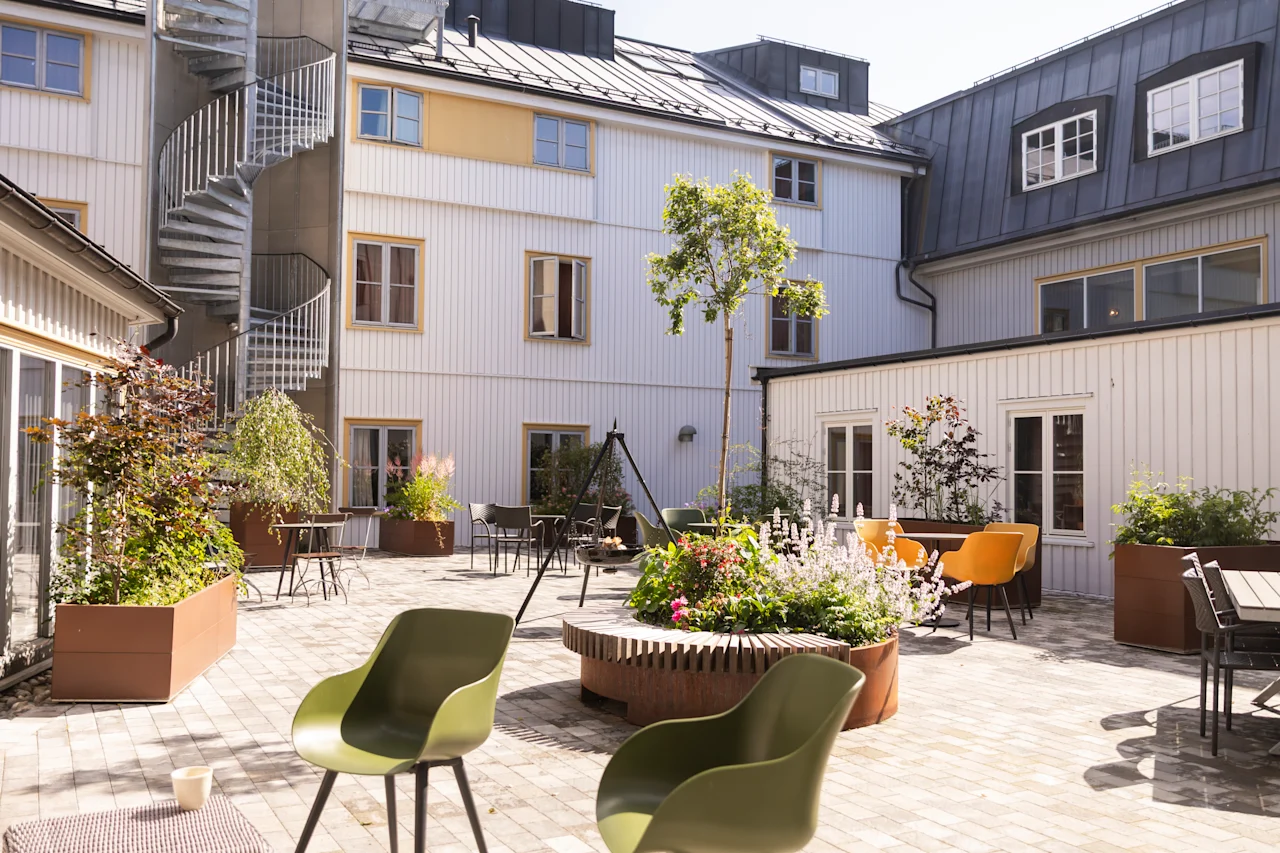 Två gröna stolar står på en solljus innergård med växter, en spiraltrappa och omgivande vita byggnader, vilket skapar en fridfull utomhusmiljö.