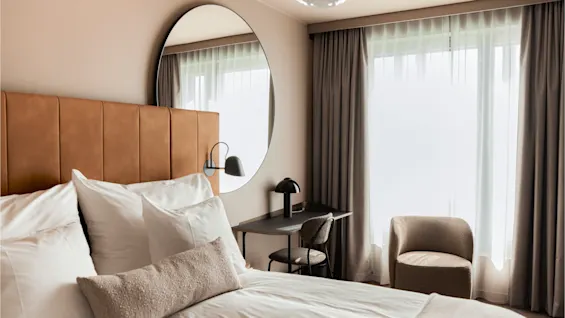 Bed with white sheets and pillows rests against a brown headboard. A round mirror, desk, and chair are adjacent. Large window with curtains brightens the room.