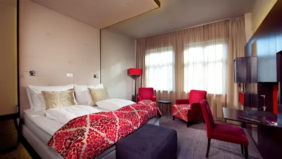 Et moderne hotellrom med pent oppredd seng, lyse røde detaljer, og naturlig lys inn gjennom et gardinbelagt vindu.