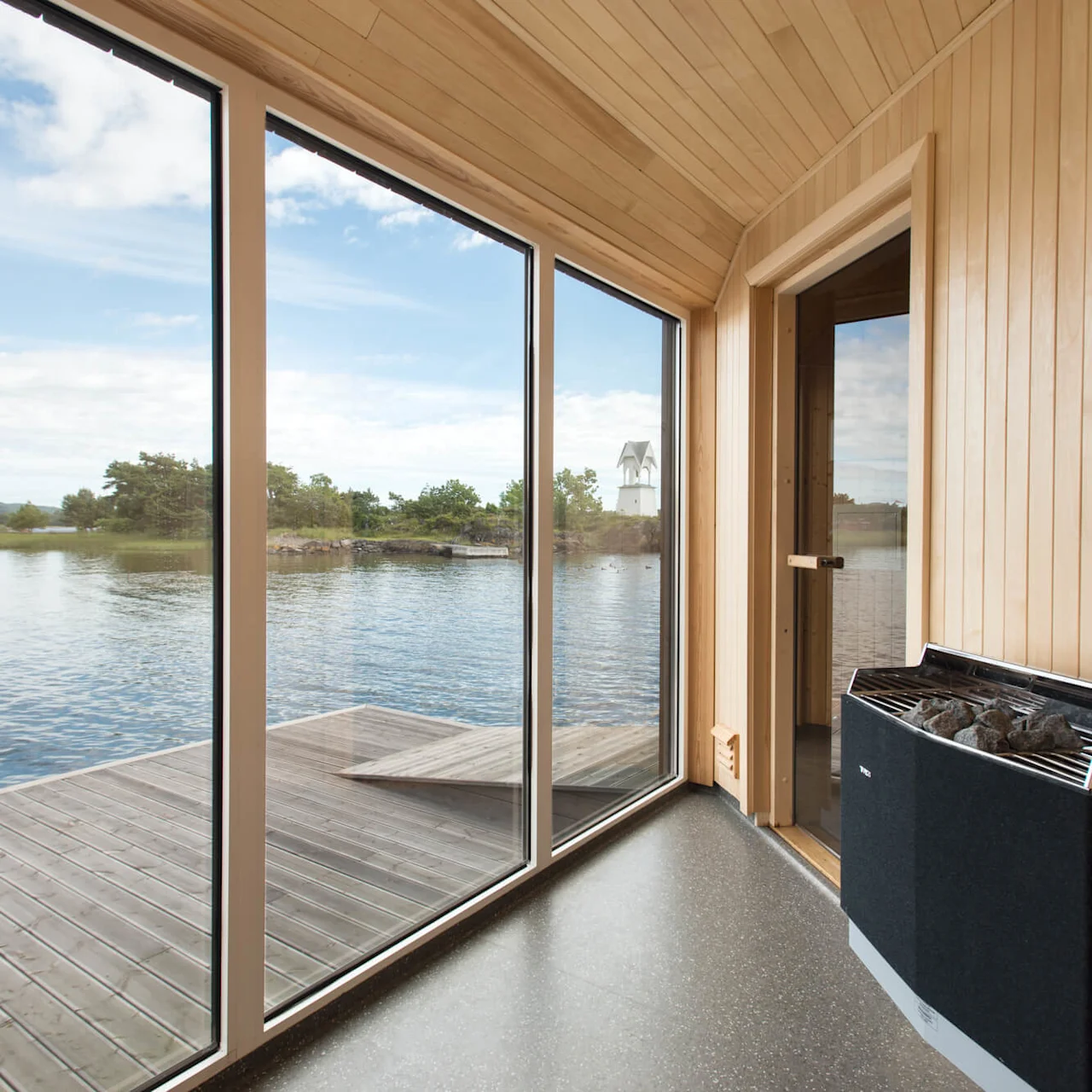 An indoor sauna features large glass windows overlooking a serene lake with trees in the distance. The wooden walls and floor create a warm, peaceful atmosphere.