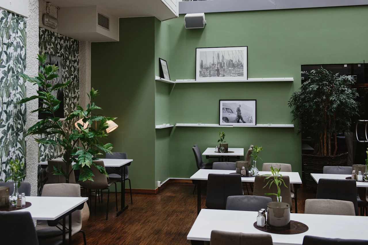 Bord och stolar är arrangerade i ett modernt, växtfyllt café med gröna väggar och inramade bilder. Miljön har trägolv, vilket ger en mysig atmosfär.