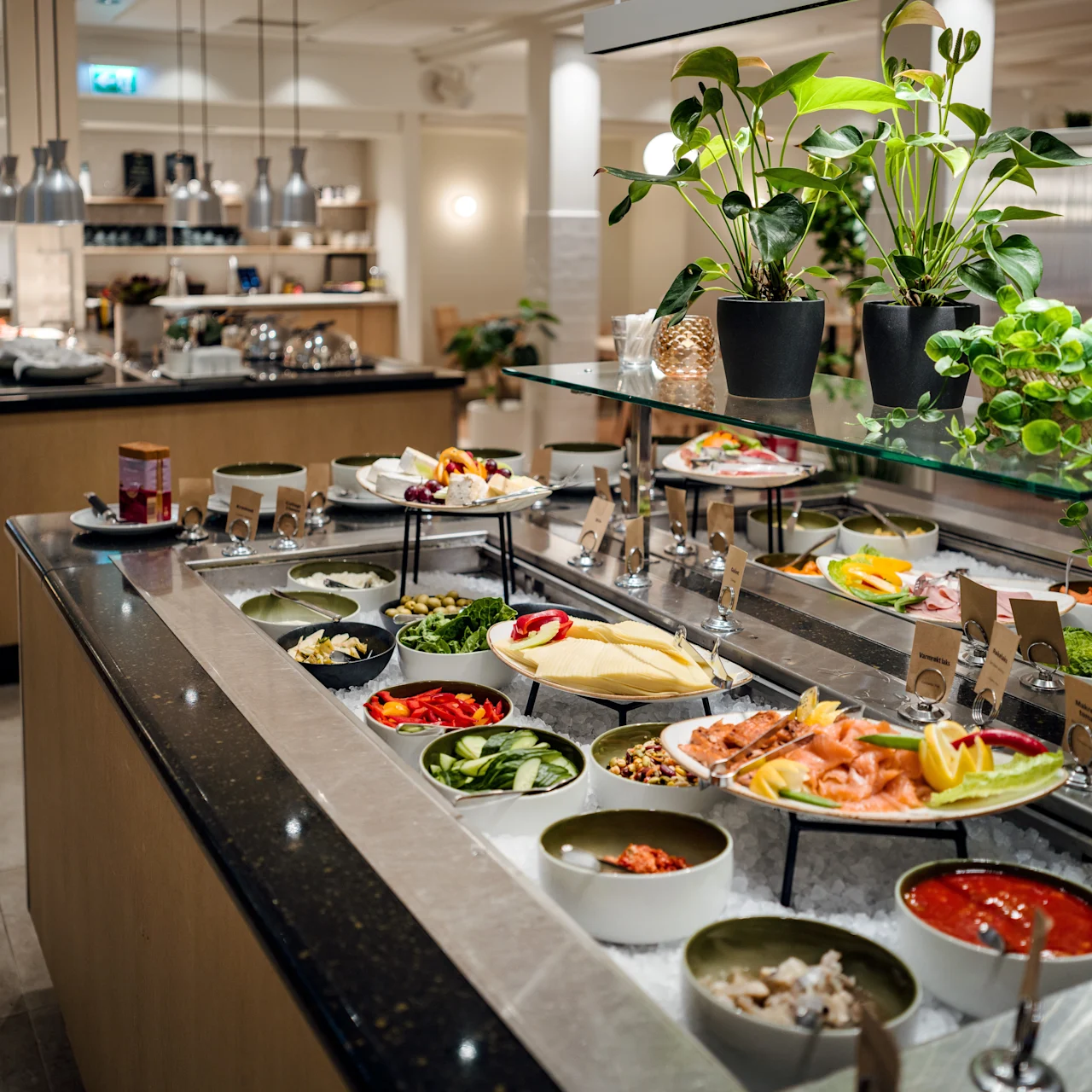 Buffet counter displays bowls of various salads, cold cuts, and garnishes. Plants decorate the upper shelf. Pendant lights hang above; a blurred kitchen area is visible in the background.