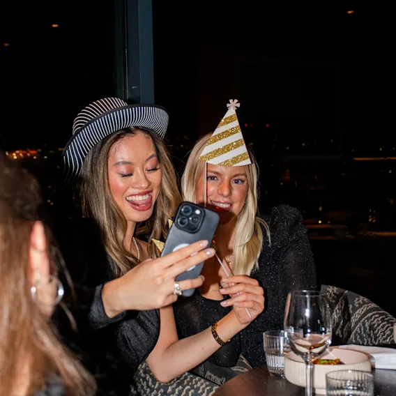 To smilende kvinner med festhatter tar en selfie med en smarttelefon ved et restaurantbord under en festlig kveld.   