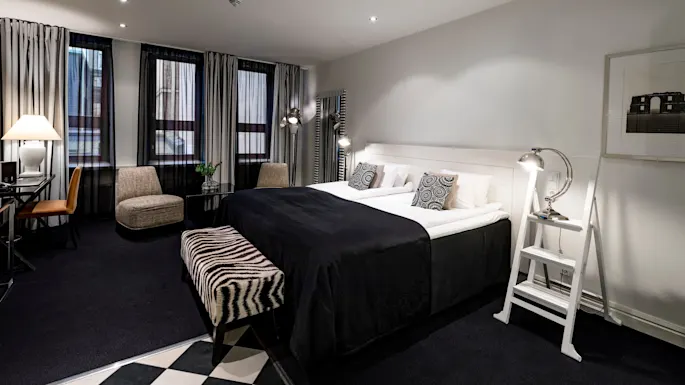 Et pent innredet hotellrom har en stor seng med svart sengetøy, mønstrete puter og en zebra-stripet benk. Moderne møbler og dekorasjoner skaper en stilig atmosfære.