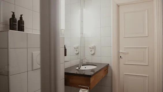 Et rent, moderne badeværelse med hvide fliser, håndvask, spejl og en hvid dør. Bekvemmeligheder er synlige på en hylde.