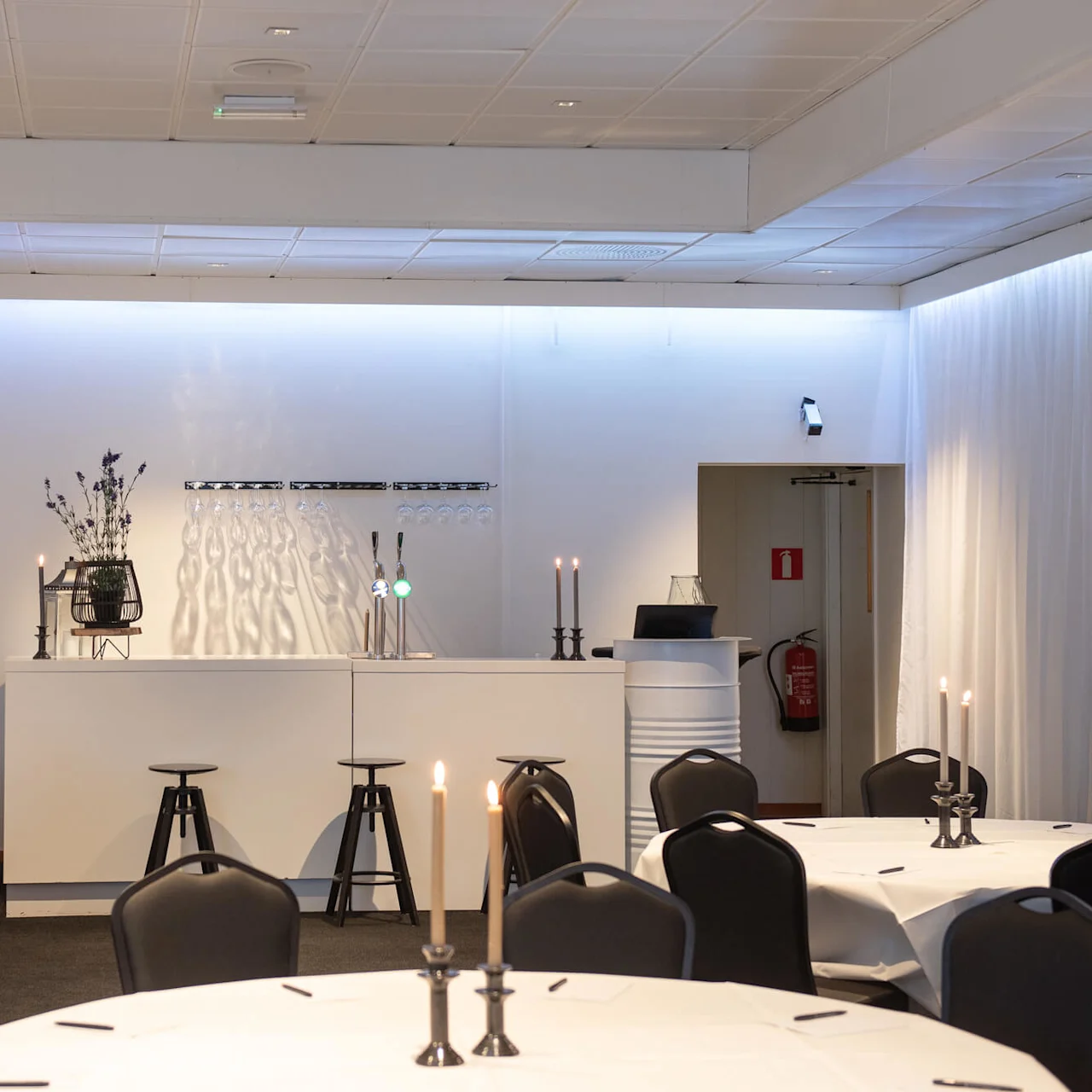 A minimalist bar area with bar stools, lit candles, and decorative flowers. Set in a softly lit banquet room with round tables draped in white tablecloths. Signage shows a fire icon.