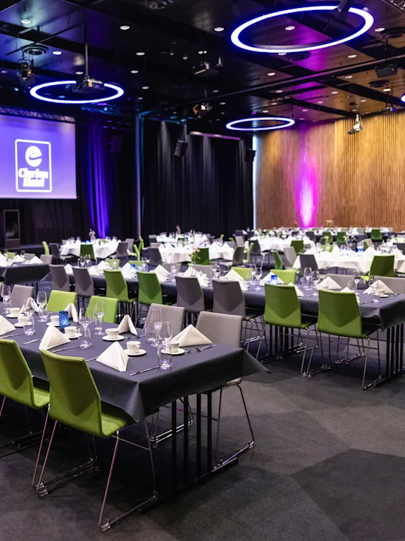 Konferensrum med rader av bord dukade med tallrikar, servetter och glas; tomma stolar omger dem. En stor skärm visar "Clarion Hotel"-logotypen. Taklampor avger ett lila sken.