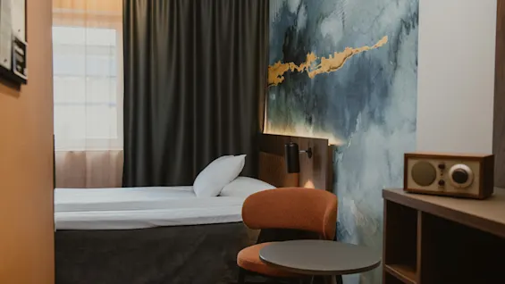 Et innbydende hotellrom med en pent oppredd seng, en komfortabel stol og en unik abstrakt kunstvegg. En radio gir et hjemmekoselig preg.
