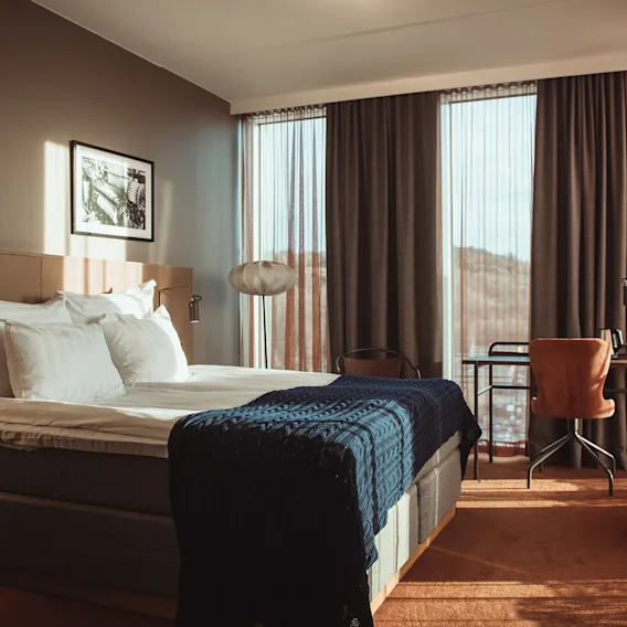 Ett ljust hotellrum med dubbelsäng, blå stickad pläd, skrivbord vid stora fönster och varmt solljus.