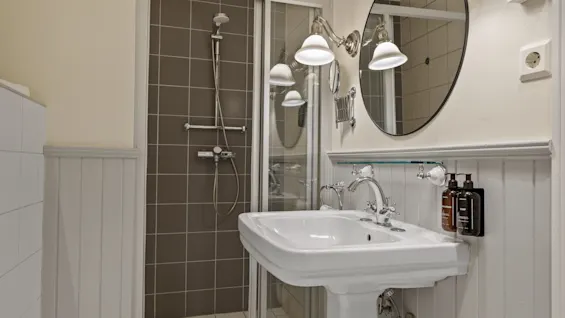 Et rent og veloplyst badeværelse med bruser med glasdøre og en piedestalvask med spejl og væglamper.