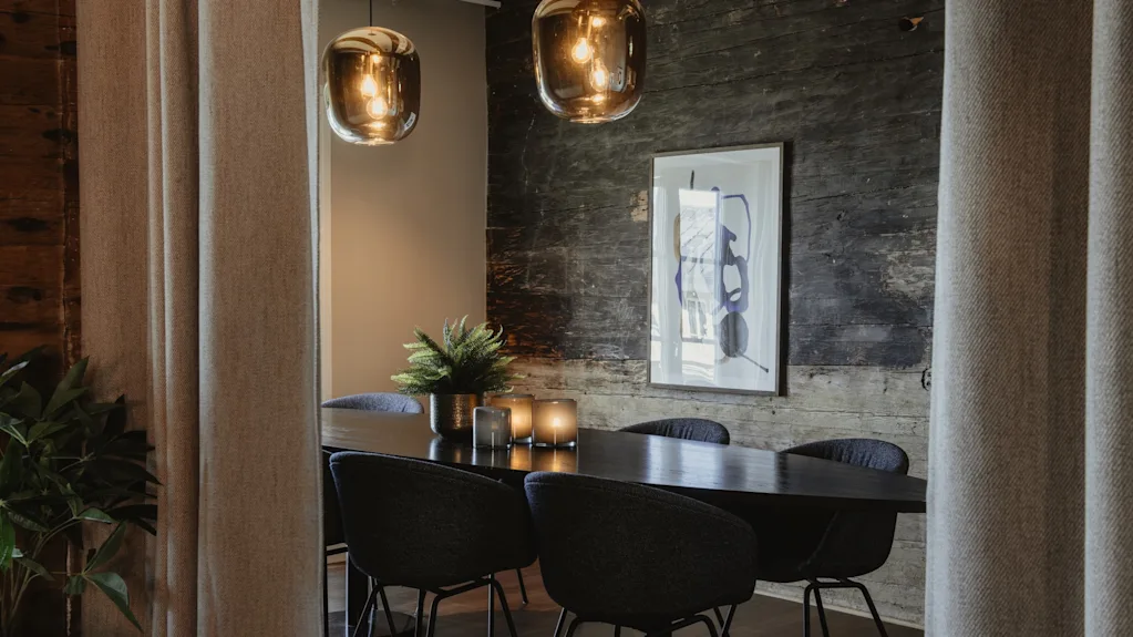Et stilfuldt interiør med et mørkt bord, moderne stole, varme pendellamper og en rustik trævæg, der skaber en indbydende atmosfære.