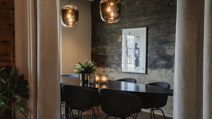 Et stilfullt interiør med et mørkt bord, moderne stoler, varme pendellamper og en rustikk trevegg, som skaper en innbydende atmosfære.