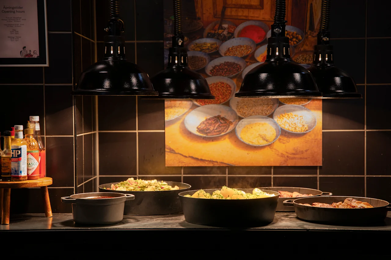 En varm buffet med varme retter i svarte serveringskar, opplyst av taklamper. Et skilt med åpningstider er synlig.