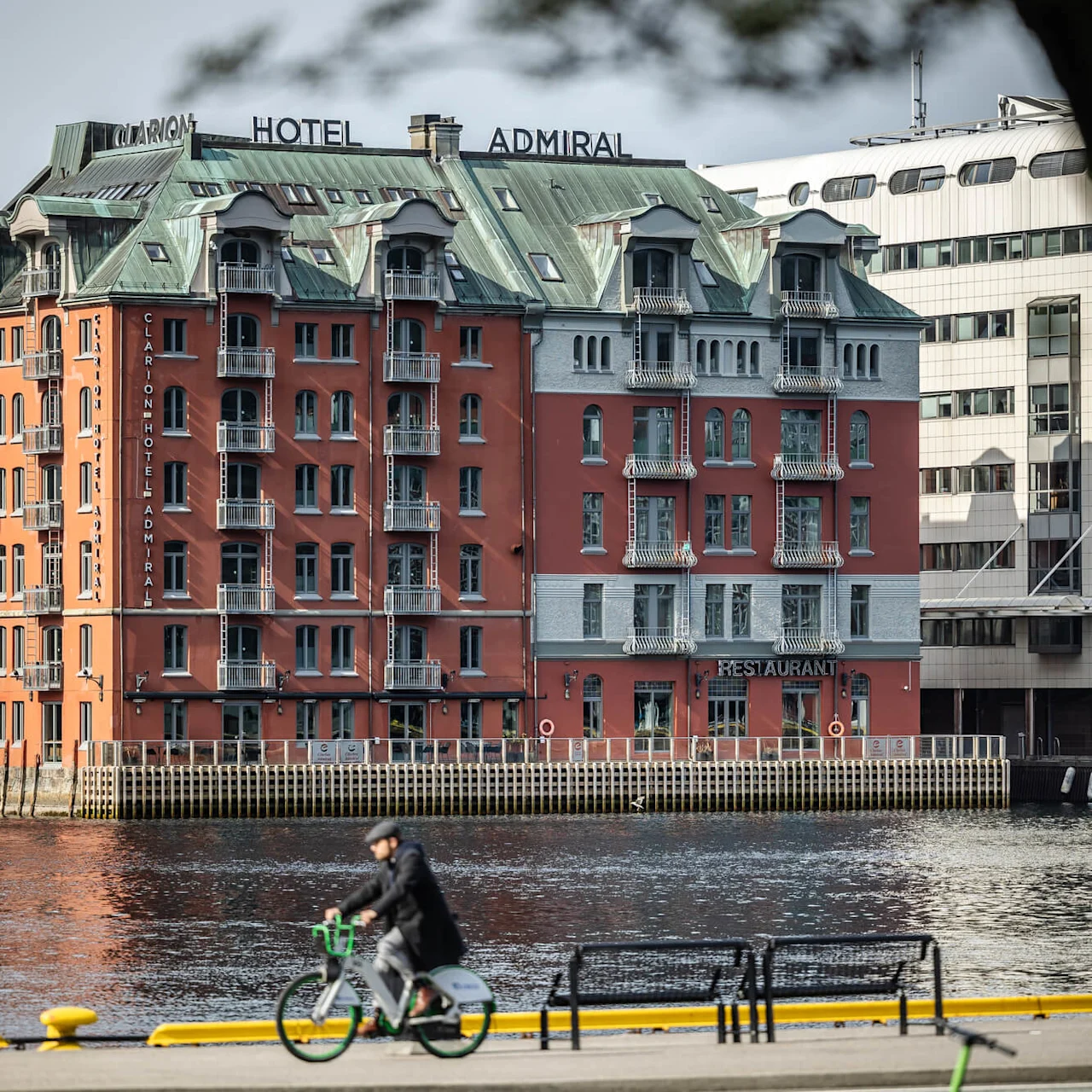Clarion Hotel Admiral, en rød murstensbygning med altaner, ser ud over en kystlinje. "RESTAURANT" skilt på facaden. En person cykler langs kajen i forgrunden.