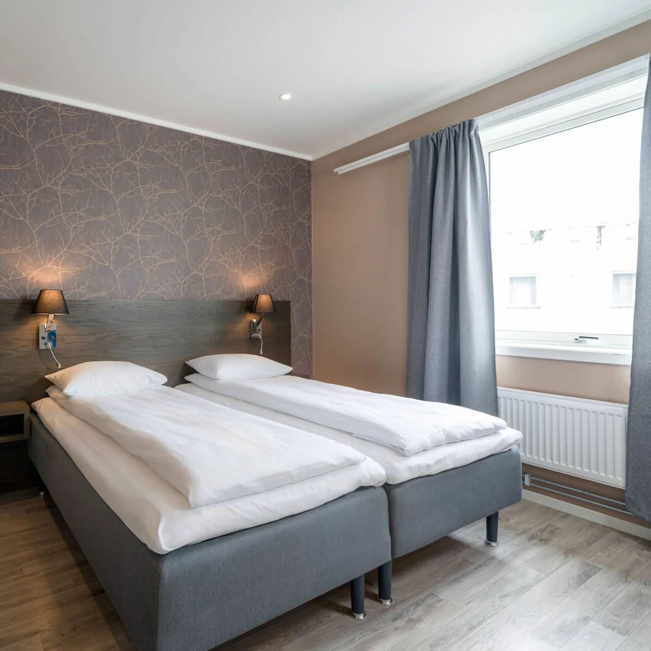 A double bed with white linens is neatly arranged against a patterned wallpaper, under soft lighting. A window with gray curtains provides natural light, set in a modern bedroom.