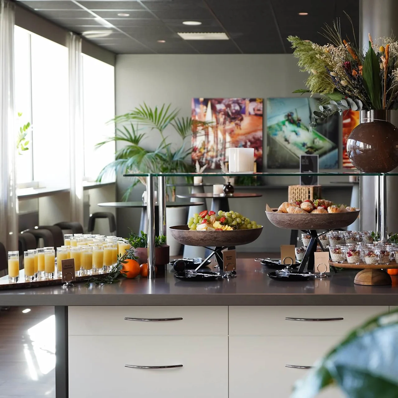 Glasses of orange juice and bowls of fruit arranged on a counter in a bright, plant-filled room with large windows.