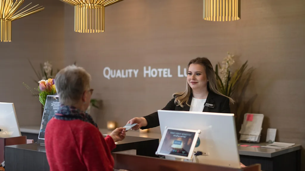 Receptionist ger ett kort till en gäst vid hotellets incheckningsdisk. Elegant belysning ovan, blommor i närheten. Skylten lyder "QUALITY HOTEL LLE.” Receptionen känns modern och välkomnande.