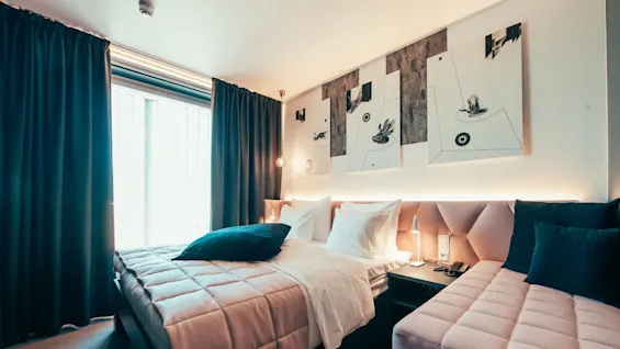 A neatly made bed with white pillows rests in a modern room, adorned with abstract wall art. Soft lighting highlights the sleek furnishings, with curtains partially covering a large window.