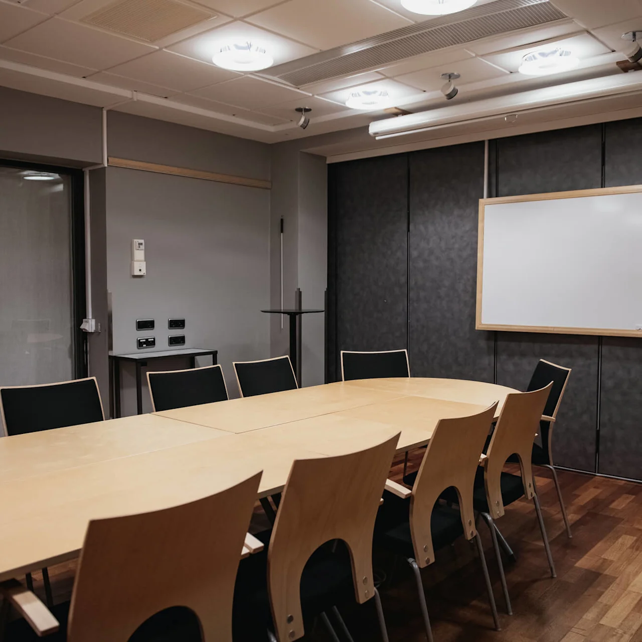 A long wooden table with chairs arranged in a conference room, under ceiling lights and a blank whiteboard on the wall.