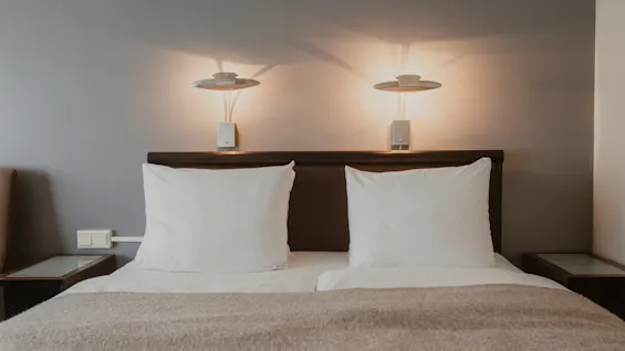 En mysig hotellsäng med vita kuddar, en mjuk filt och varm belysning från två vägglampor ovanför sänggaveln.