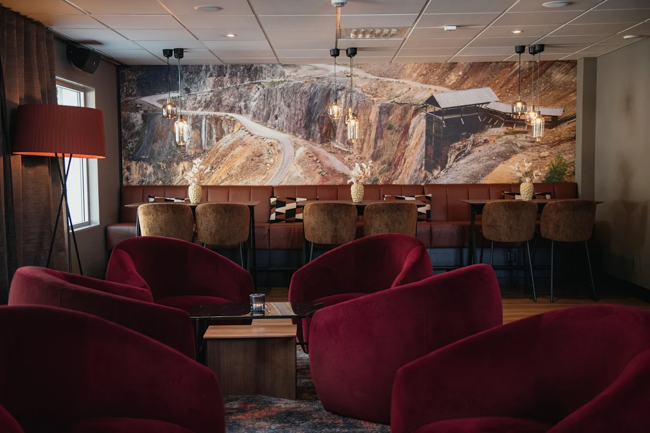 Et komfortabelt salongområde med myke røde lenestoler og benkeseter, med et stort veggmaleri av et ulendt landskap.