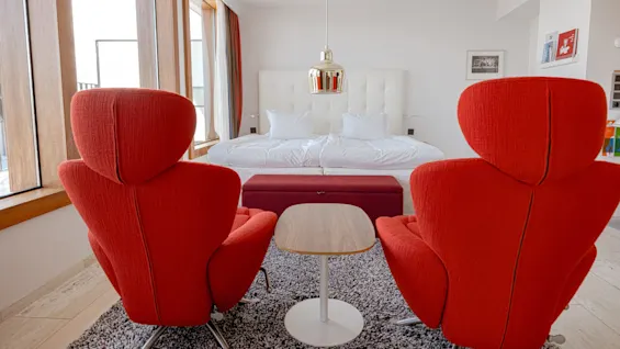 Two red chairs face a wooden table in a modern bedroom with large windows, white bedding, and a pendant light above, creating a cozy and stylish atmosphere.