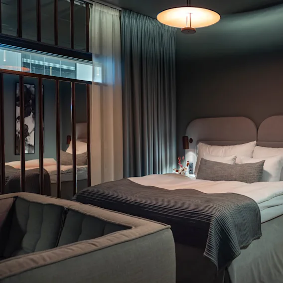 En seng med hvidt og gråt sengetøj er pænt arrangeret, placeret i et svagt oplyst, moderne soveværelse med reflekterende overflader, gardiner og en hyggelig atmosfære.