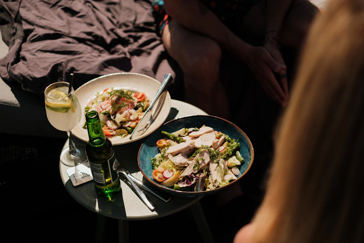 Två skålar med färsk sallad står på ett litet utomhusbord, tillsammans med en citroncocktail och en grön flaskdryck. Människor sitter i närheten i soligt väder.