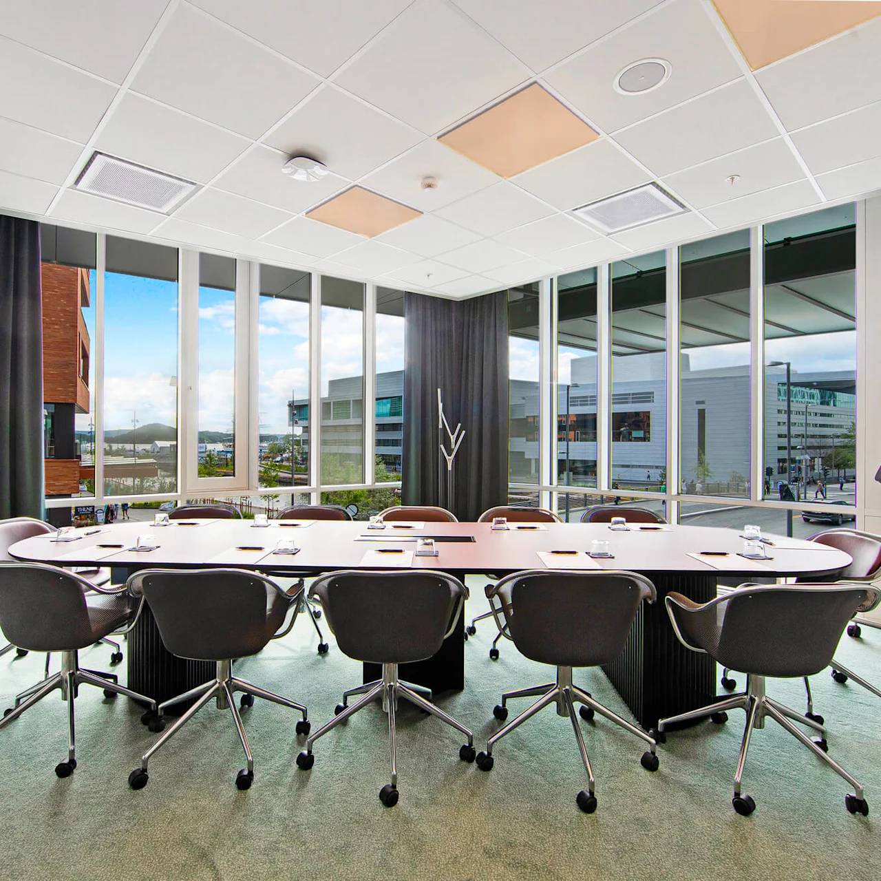 A conference room features a long table surrounded by swivel chairs. Through large windows, a modern building complex is visible. A flip chart displays "Clarion Hotel Oslo."