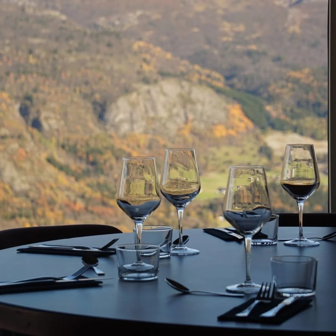 A round dining table set with wine glasses, utensils, and plates, overlooking a scenic mountainous landscape with autumn foliage.