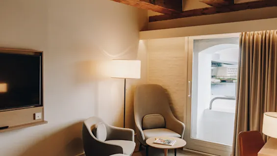 Ett mysigt hotellrum med fåtöljer, TV och balkong med utsikt över vatten. Synliga takbjälkar.