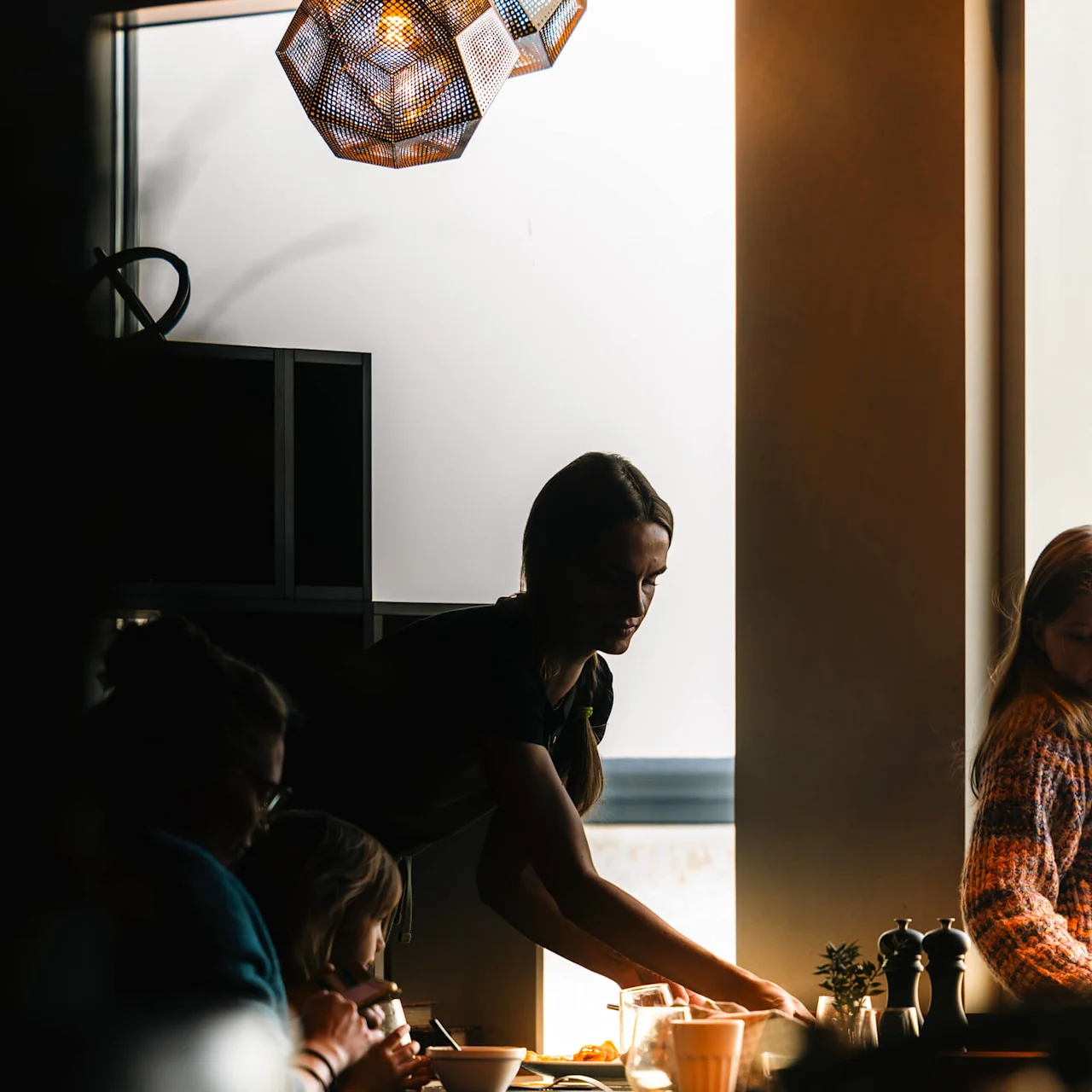 Servitris serverar ett bord med drycker och rätter på ett svagt upplyst kafé. Personer sitter i närheten, upplysta av en geometrisk ljusarmatur.