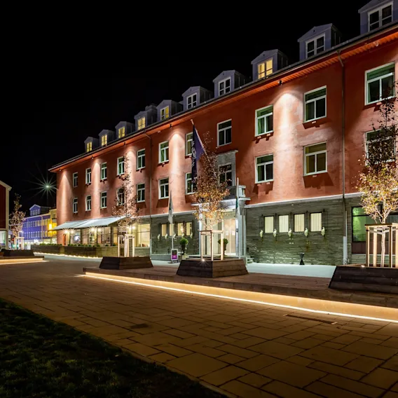 En godt opplyst bygning om natten, muligens et hotell, med trær i plantekasser og opplyste stier som fører til inngangen.