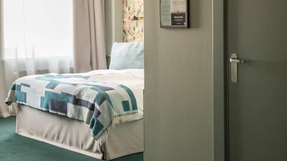 En prydligt bäddad säng med ett mönstrat täcke står i ett rum med blommigt tapet och ett stort fönster, omgiven av mjukt ljus och grön matta. En vägg har hotellinformation.