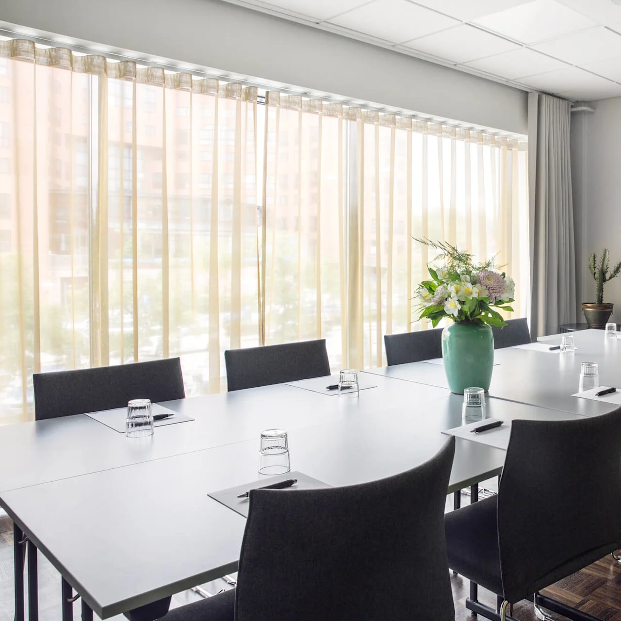 Ett konferensbord är dukat med anteckningsblock och glas, omgivet av stolar. En vas med blommor pryder mitten. Solljus filtreras genom skira gardiner bredvid en whiteboard.
