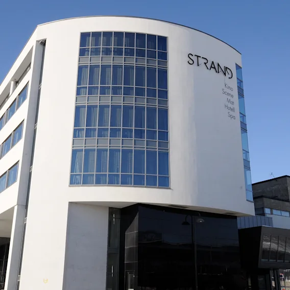 En moderne fleretasjes bygning med store glassvinduer buer oppover under en klar blå himmel. "STRAND" er tydelig vist på fasaden med mindre tekst: "Sola Hotel Spa."