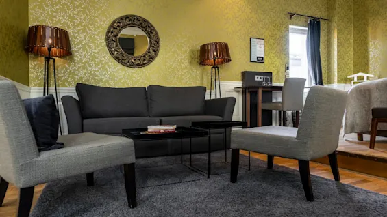 A gray sofa sits between two chairs on a rug, accompanied by a round mirror and floor lamps against a yellow patterned wall, with a desk nearby.