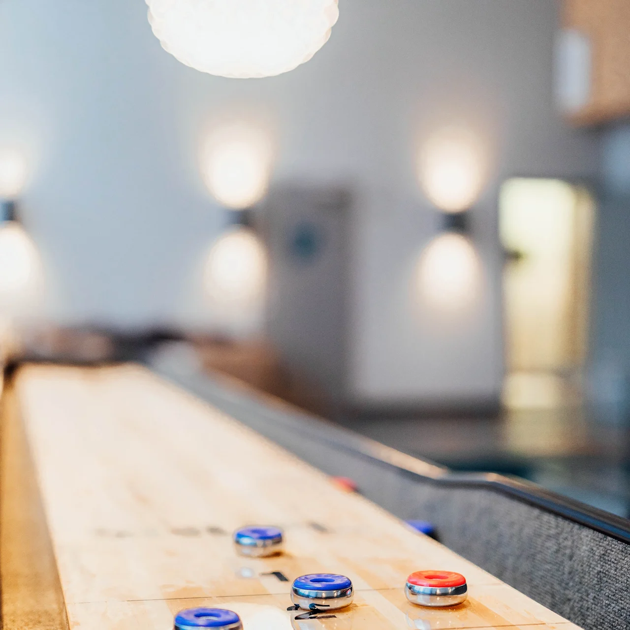 Shuffleboardstycken ligger på ett träbord, redo för spel. En ljus, sfärisk taklampa lyser upp rummet med suddiga vägglampor i bakgrunden.