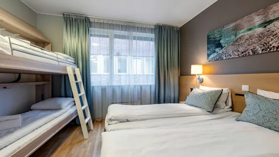 Two single beds and a bunk bed are in a tidy bedroom with wooden floors. A window with curtains lets in daylight, and a wall lamp provides indoor lighting.