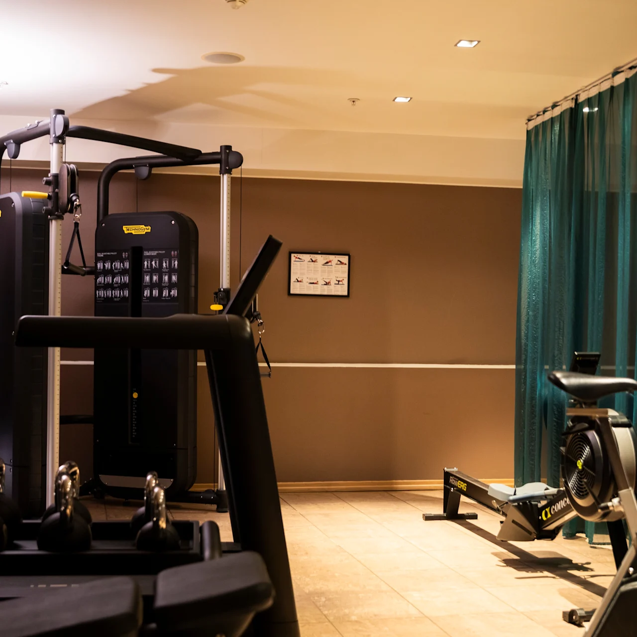 A gym with exercise machines, kettlebells, and a stationary bike in a well-lit, brown-walled room. Equipment is organized; a teal curtain partially separates areas.