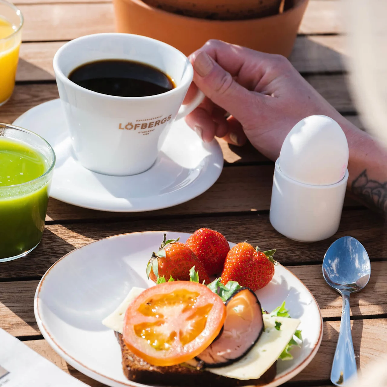 En hånd holder en hvid kaffekop med teksten "Löfbergs", ved siden af glas med appelsin- og grøn juice på et træbord. En tallerken med sandwich, tomater, jordbær og en avis er i nærheden.