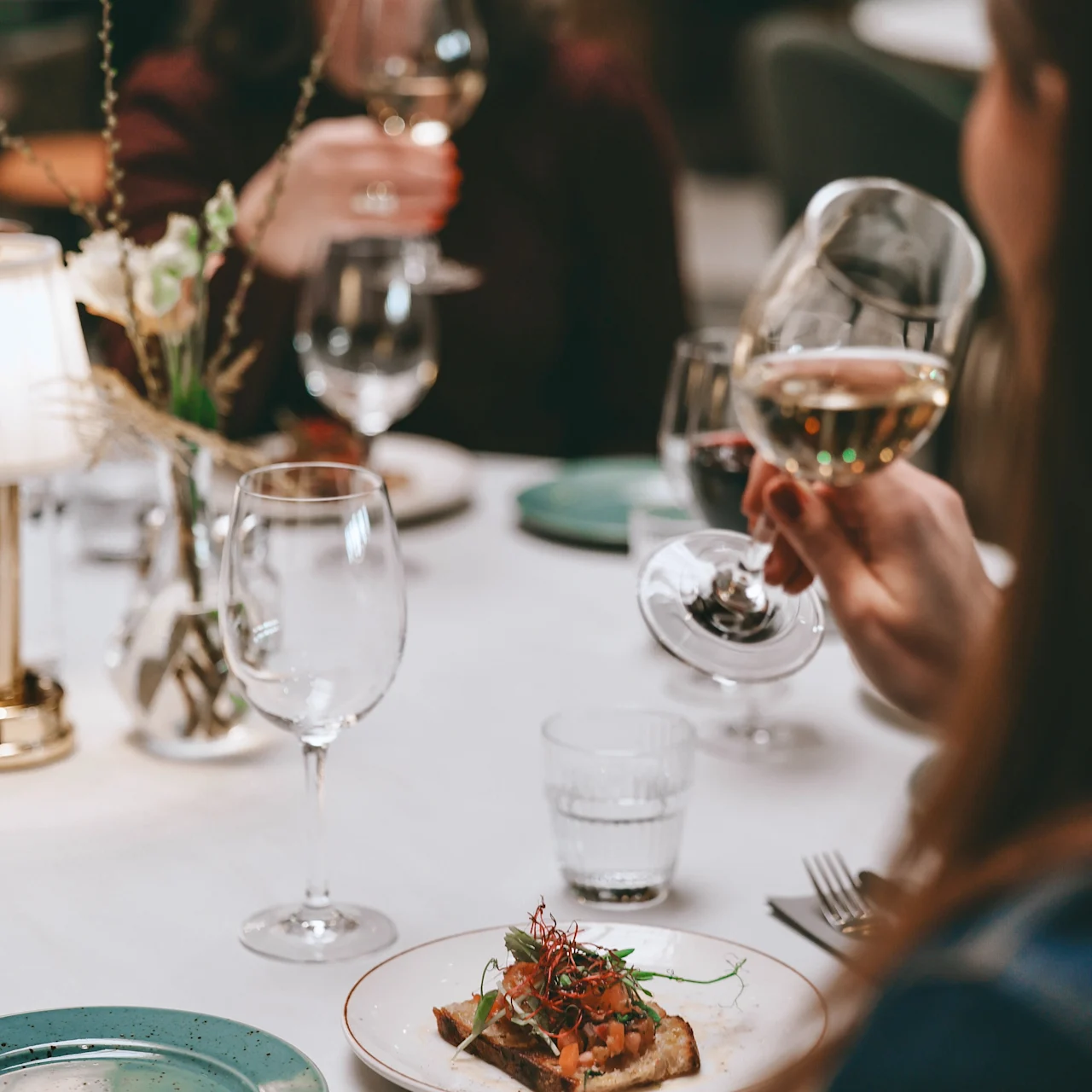 A plate with toast topped with greens sits on a table. A person holds a wine glass, with other diners blurred in the background, suggesting a social meal setting.