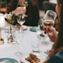 A plate with toast topped with greens sits on a table. A person holds a wine glass, with other diners blurred in the background, suggesting a social meal setting.