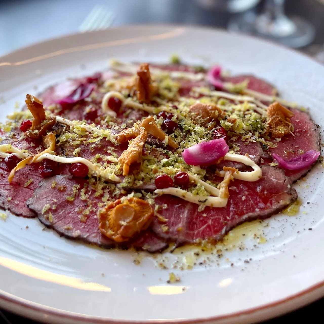 Sliced beef carpaccio topped with mushrooms, pink onions, berries, grated garnish, and drizzled with sauce on a large white plate; set on a tabletop with blurred background.