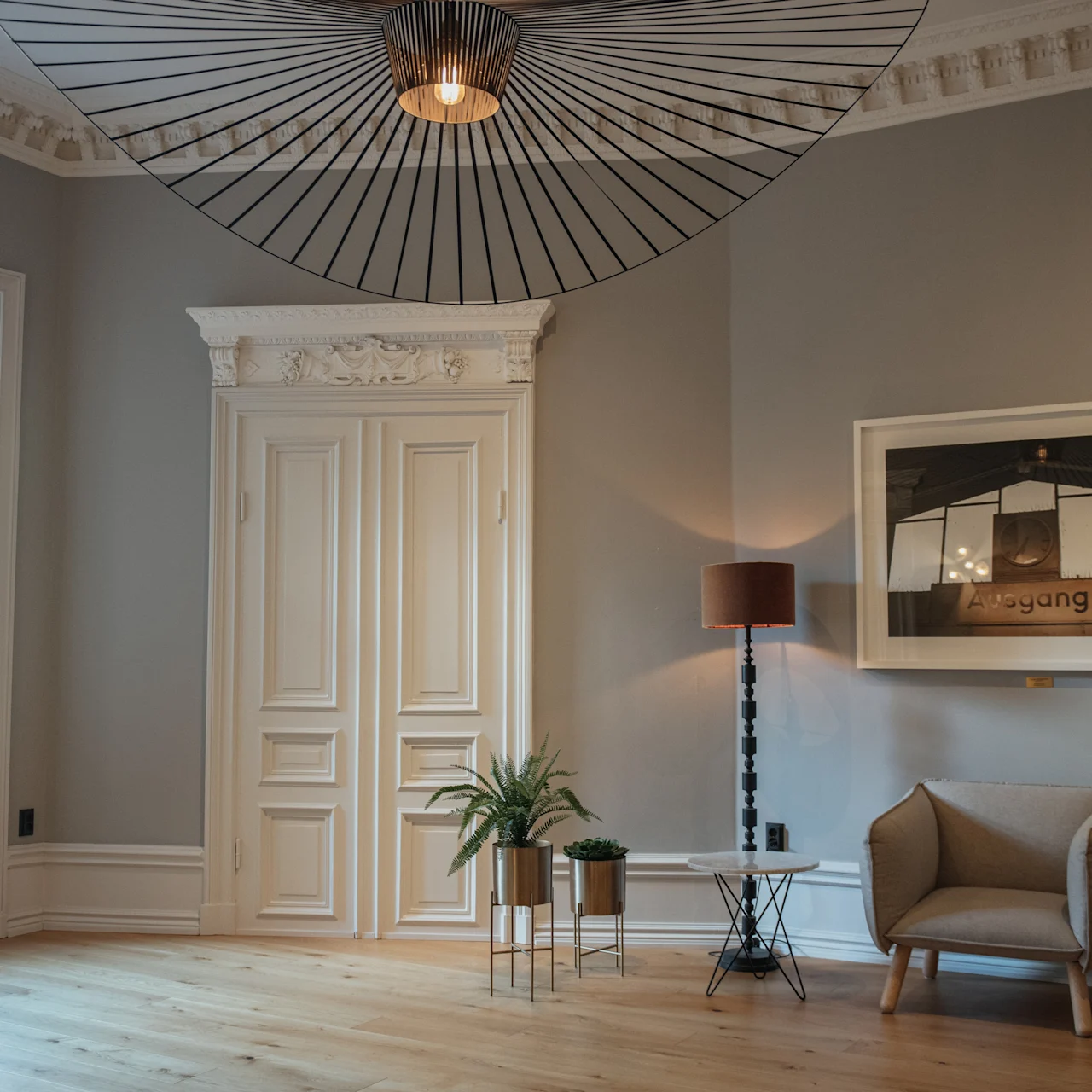 Ett stilfullt vardagsrum har en modern fåtölj, en dekorativ golvlampa och krukväxter. Ett stort inramat fotografi pryder väggen. Solljus strömmar in genom ett fönster.